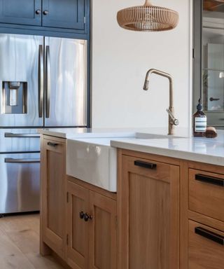 A modern rustic kitchen with wooden cabinetry, a Belfast sink, and black hardware. Rattan pendant lights and exposed beams complement the warm, airy design.