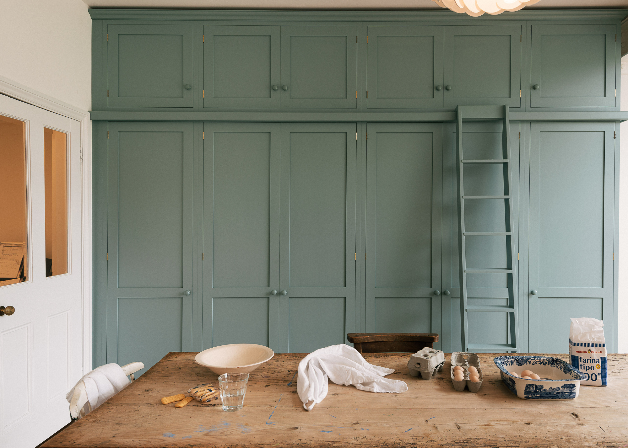 A traditional light turquoise run of floor to ceiling kitchen cabinetry in shaker style along the back wall with a matching ladder to reach the high cupboards.