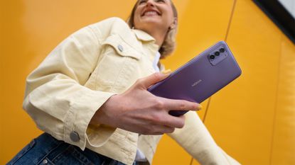The Nokia G42 5G in So Purple, held by a person stood against a yellow background