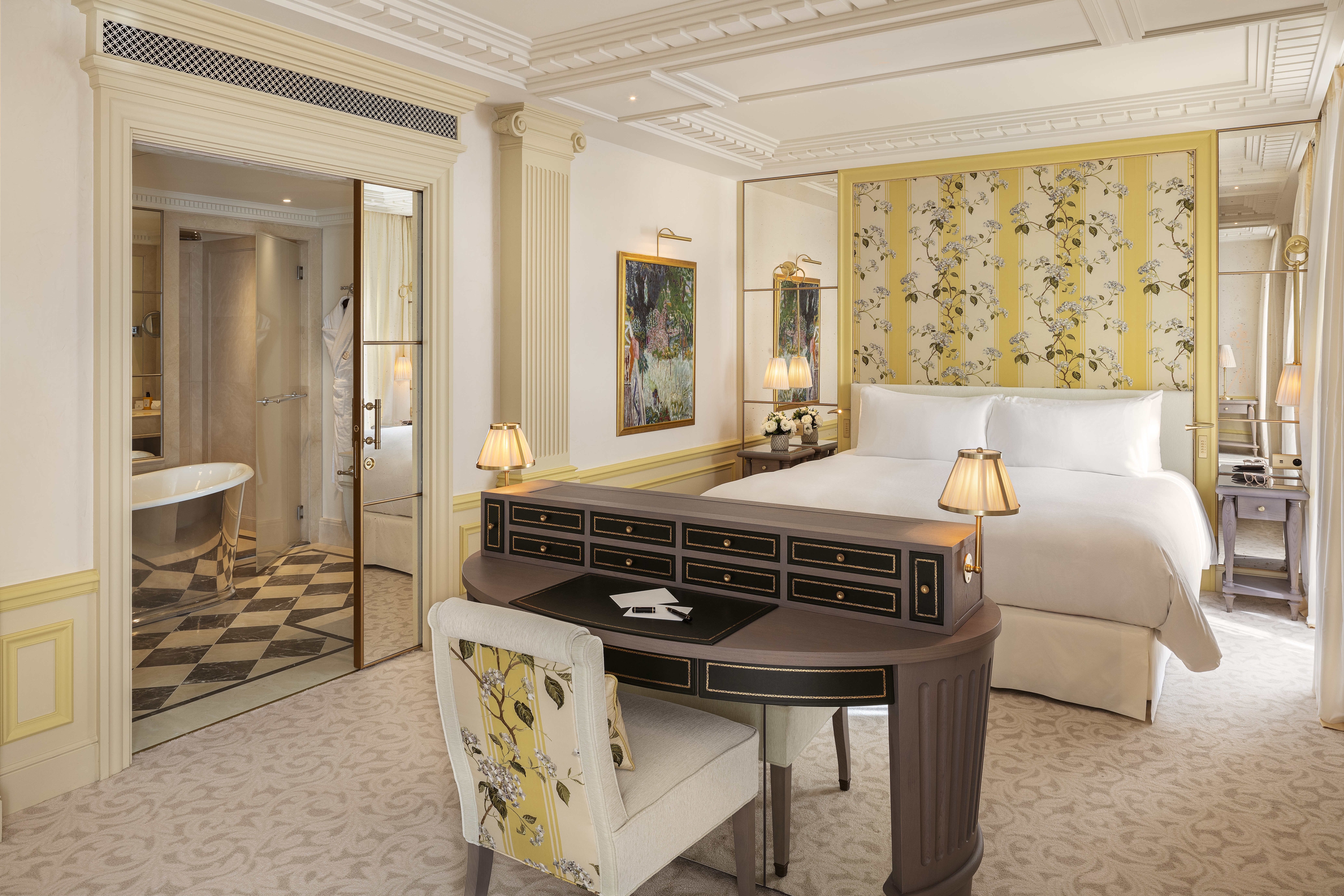 Hotel bedroom featuring a upolstered bed with white linen, a pale brown dressing table and pale yellow walls with floral detailing