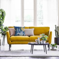 A mustard yellow velvet two-seater sofa with tall legs in a brightly lit living room