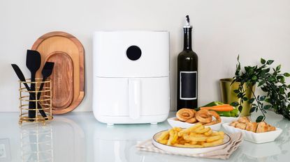 A white air fryer in a white kitchen, with an oil bottle and some plates of fried food