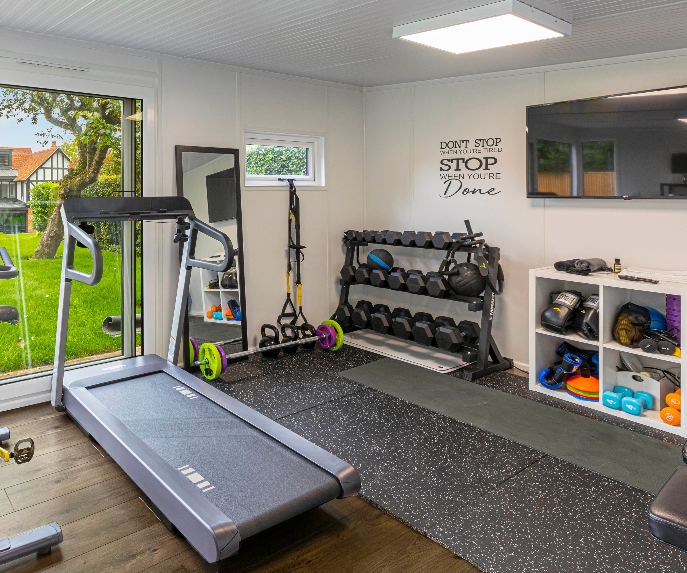 Home gym with treadmill, dumbbell rack, weights on speckled rubber flooring, garden view through doors.