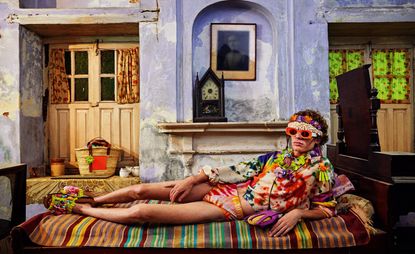 Guy in colourful clothing lying on colourful bed with colourful room surroundings