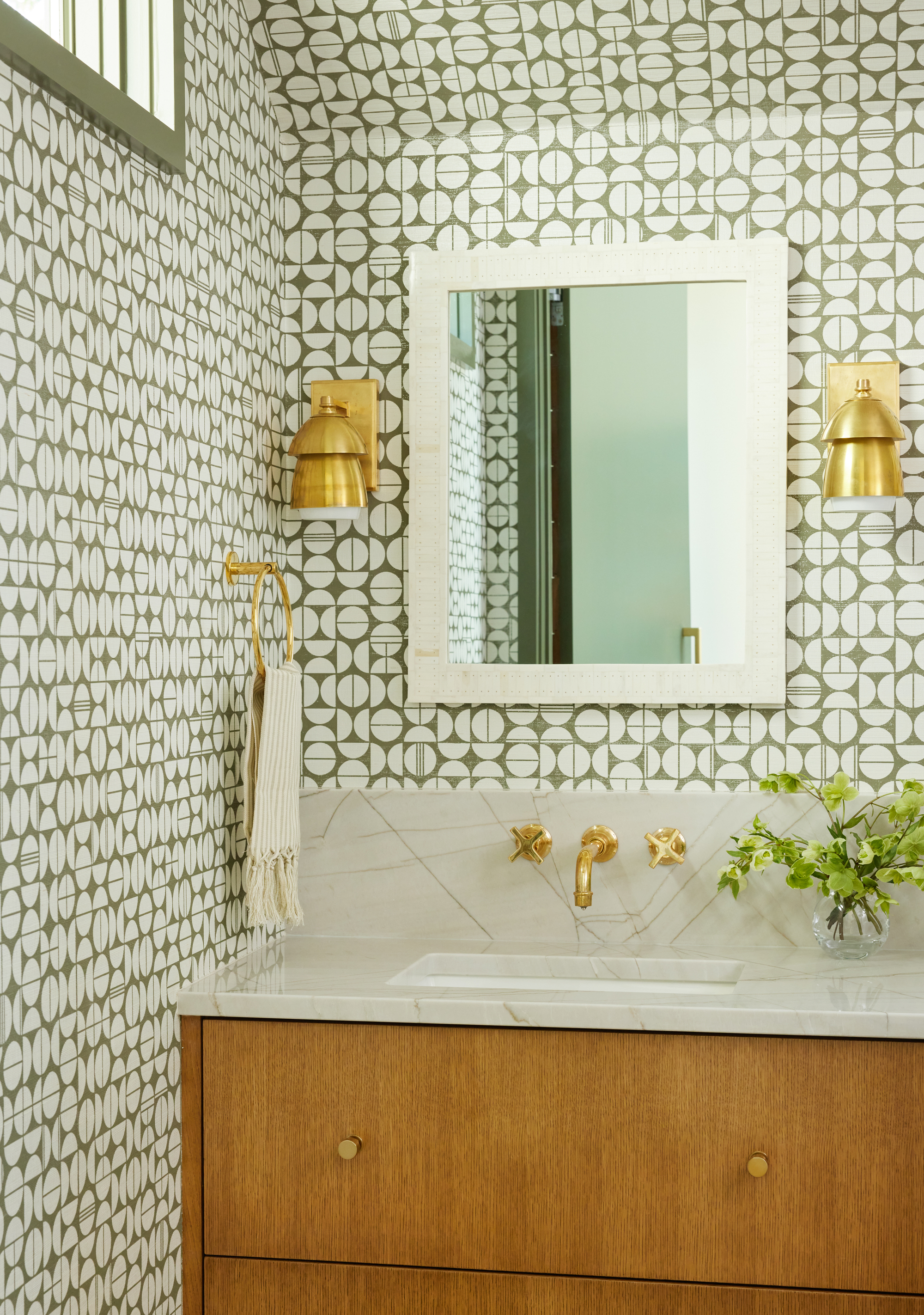 mid century modern bathroom with patterned green and white wallpaper all over, a timber vanity with white stone counter, gold taps, gold wall sconces, and a vase with flowers