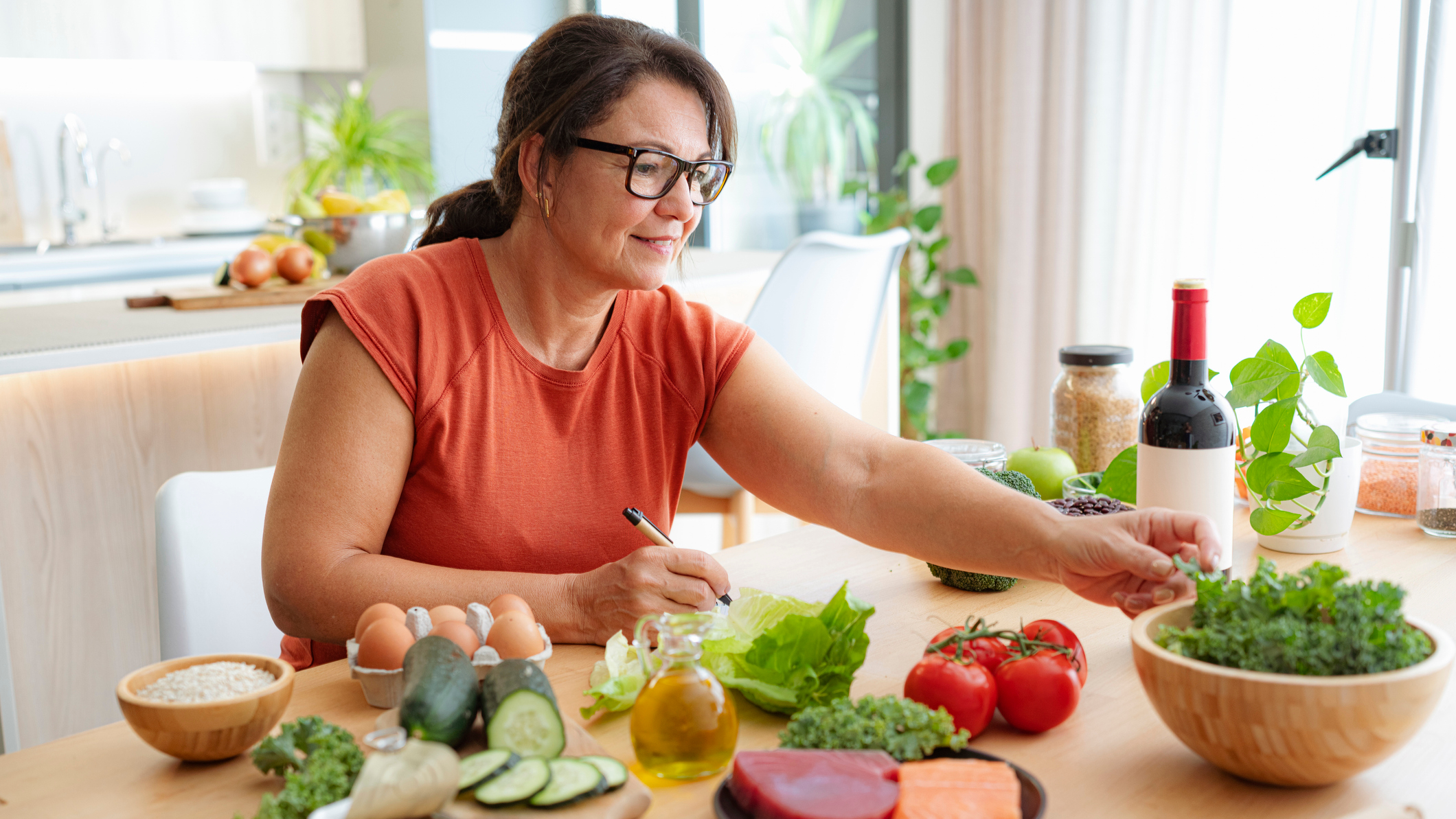 Woman sitting at a kitchen table surrounded by whole foods, including salad, beef, salmon, eggs and grains.