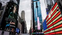 A photo of Time Square with a rocket shown on a light-up display. On the right is a light-up American Flag and on the left is a giant screen showing Elon Musk above the Nasdaq sign.