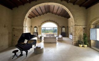 A hotel lounge area with 4 white sofas, 2 wooden consoles with a mirror on, a piano, potted plants, light floor tiles and stone walls.