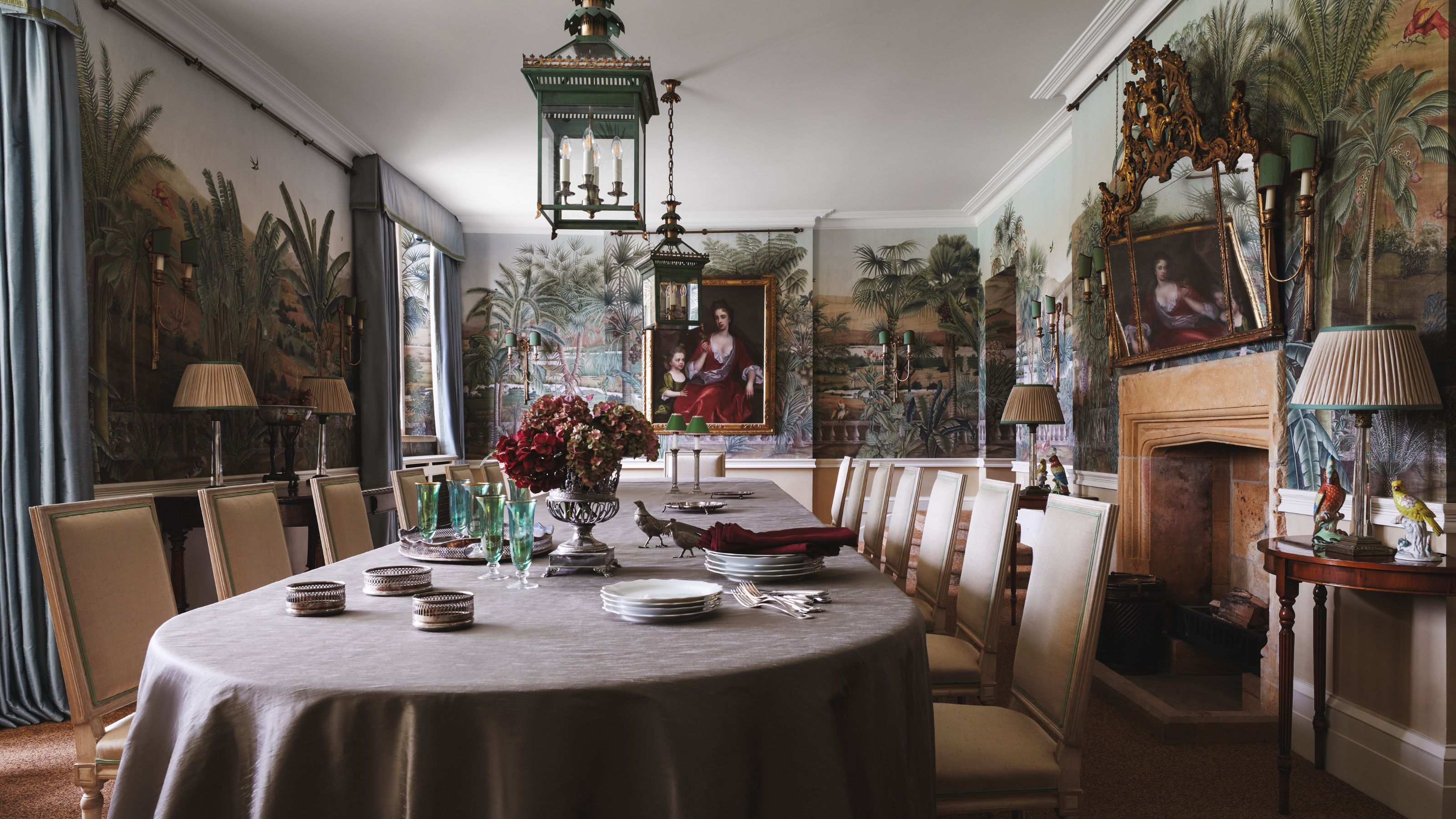 a grand dining room with a mural wallpaper, antique artwork, a gold git mirror, fireplace, and a large dining table