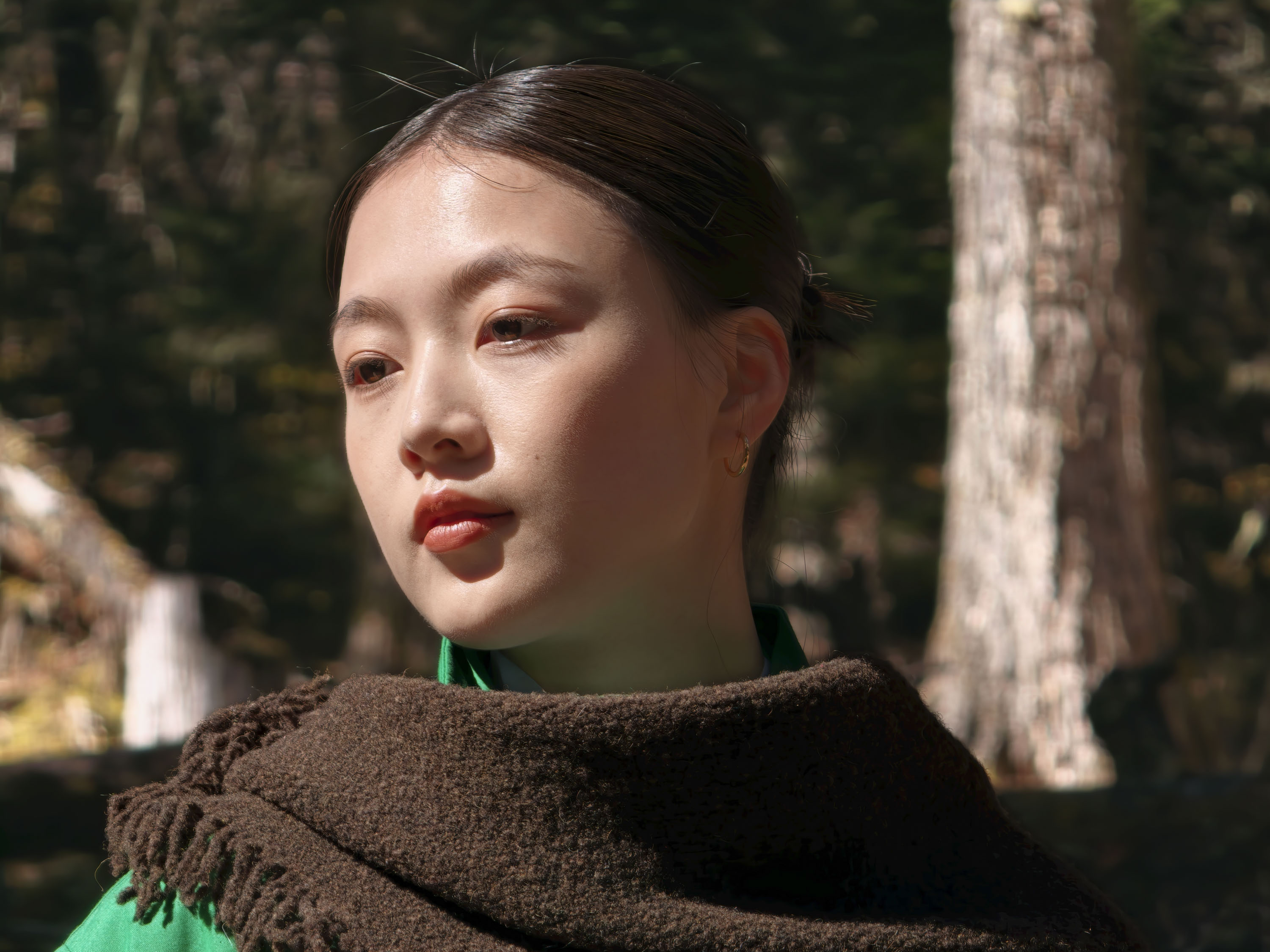 Portrait of a woman in a shawl and green jacket in a woodland, there's direct sunlight on her
