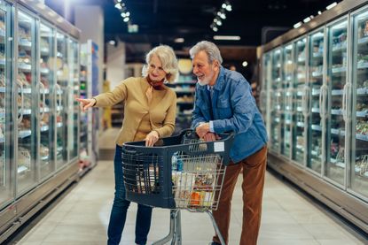 A senior couple stands in the aisle of a grocery store looking at groceries together.