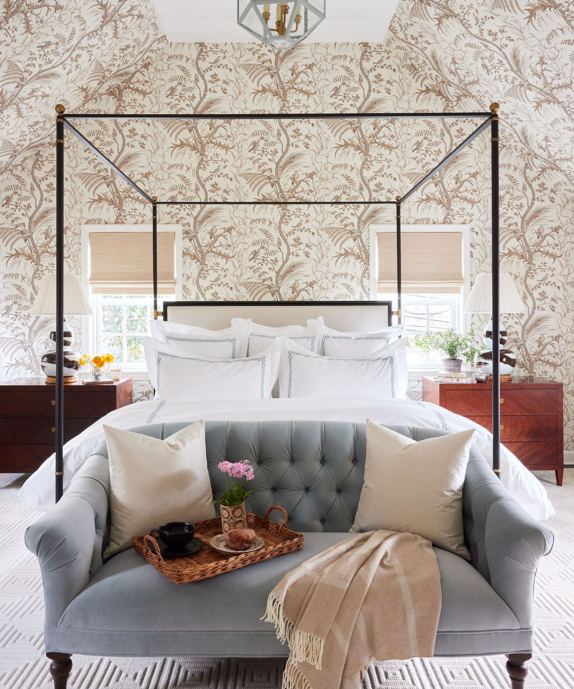 Bedroom with floral neutral wallpaper, a black four poster metal bed, hotel bedding, mahogany nightstands, and a powder blue loveseat at the end of the bed