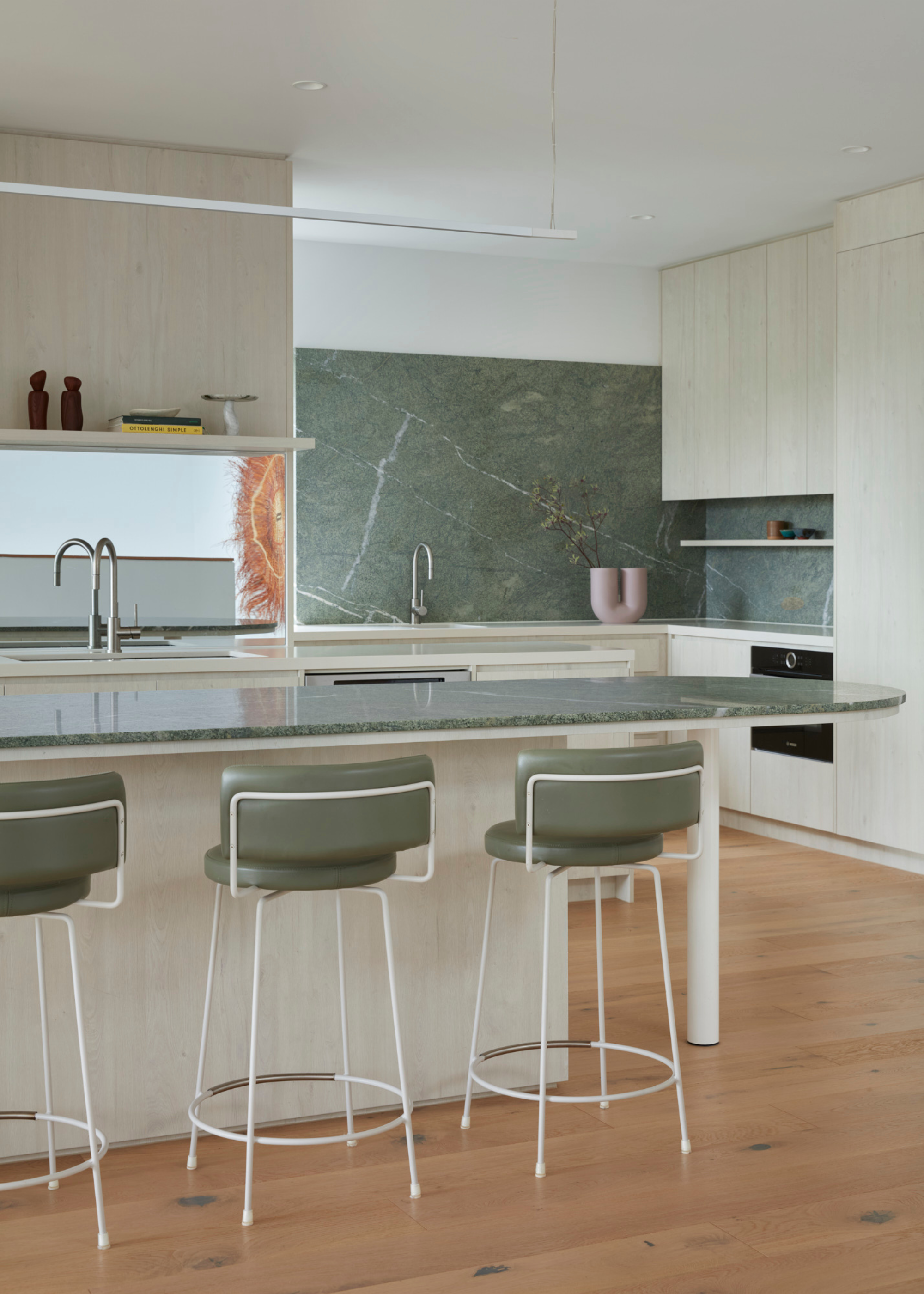 A kitchen with a green stone counter, green and white bar stools, a curved vase and silver hardware