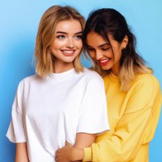 Two young women standing and cuddling, with bright blue background