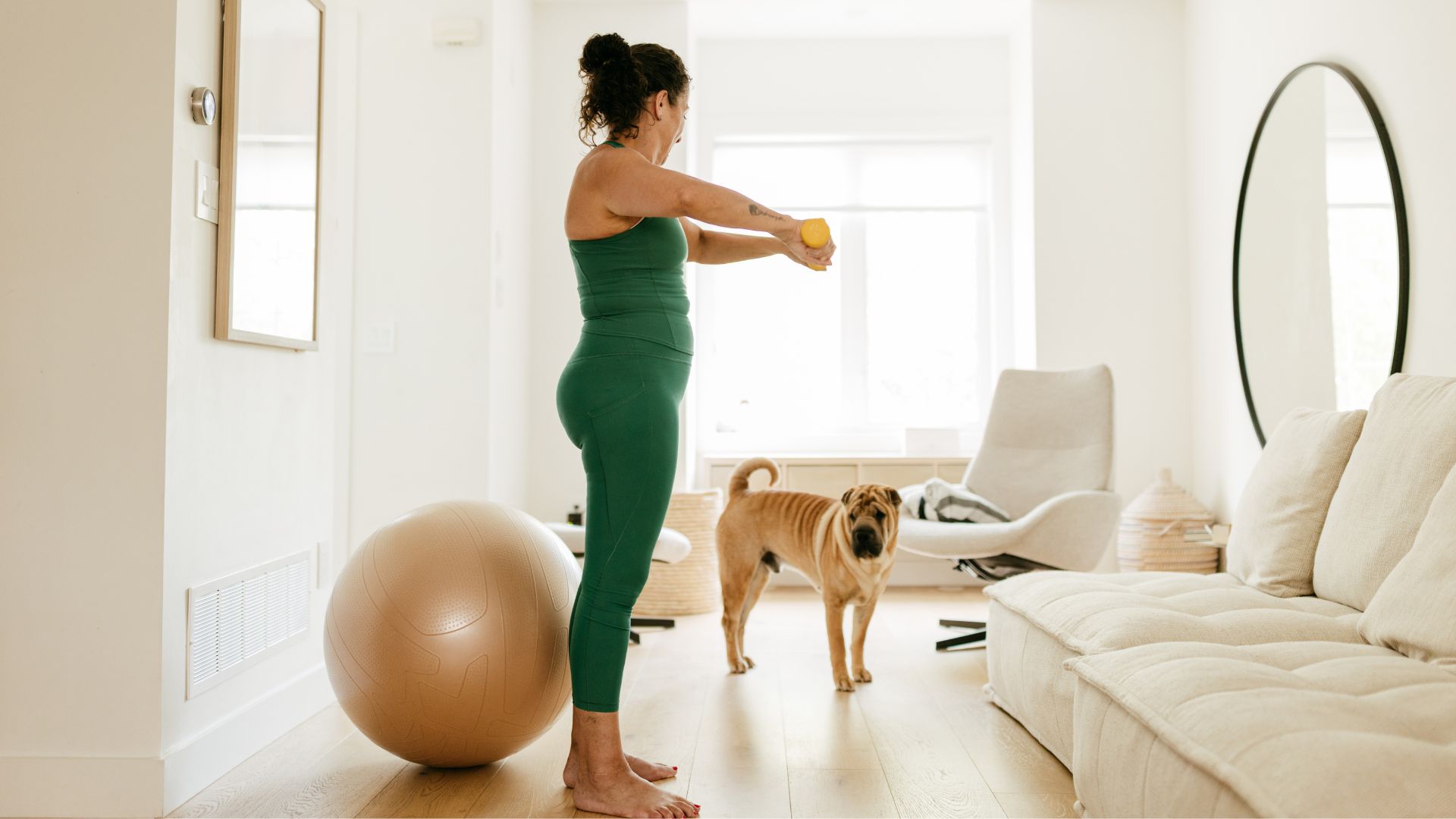 Woman standing in living room holding two light dumbbells next to bosu ball and dog