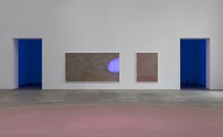 Two paintings on a white wall, between two entrances to a blue-lit room