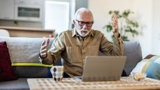 An older man throws his hands in the air with frustration as he sits on his sofa and looks at his laptop.