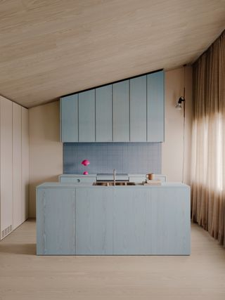 Image of a minimalist kitchen design. The cabinets are stained in a light blue color over natural wood. The backsplash is also a light blue tile, and there is a hot pink, mini mushroom lamp on the counter.