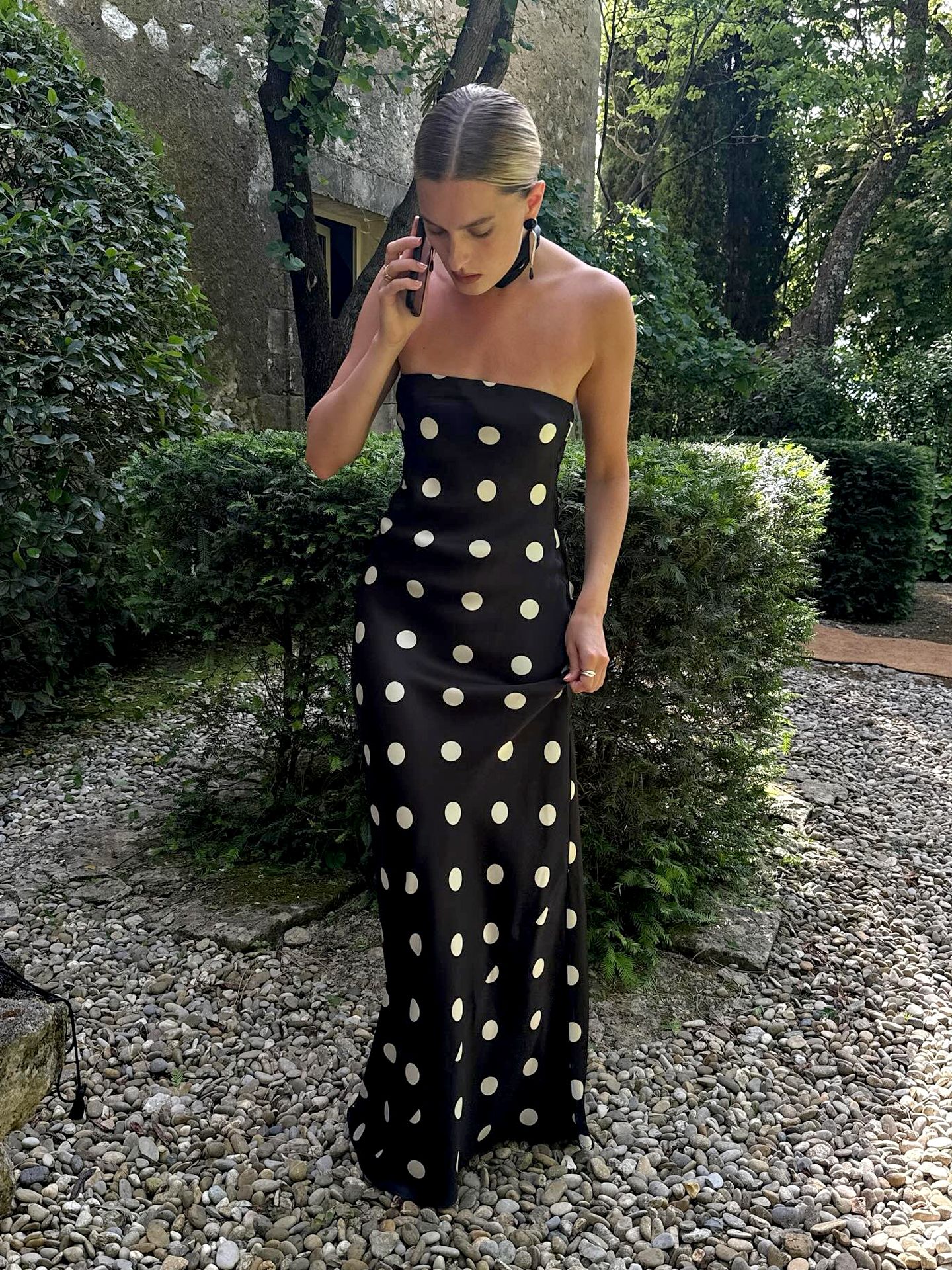 NYC style influencer Christie Tyler standing in a garden while on her phone in Provence, France wearing a polka-dot Oren Silk Dress with neck sacrf
