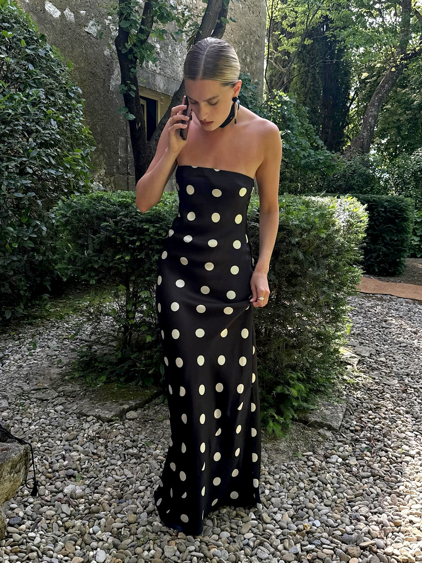 NYC style influencer Christie Tyler standing in a garden while on her phone in Provence, France wearing a polka-dot Oren Silk Dress with neck sacrf