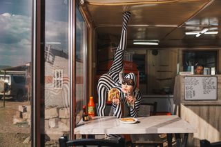 A person in a striped outfit performs a leg extension while holding food and drinks at a table in a casual eatery