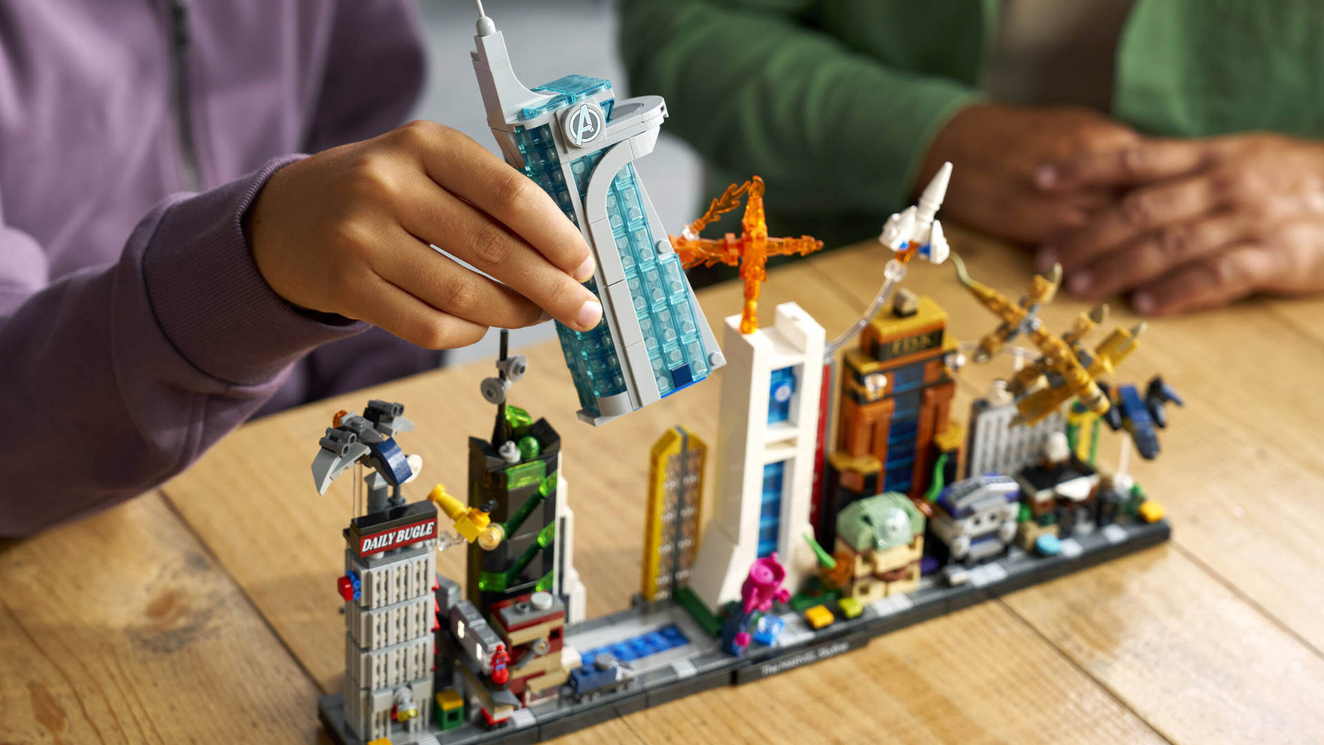 A hand holds the Avengers Tower from Lego Marvel Skyline, which sits on a wooden table