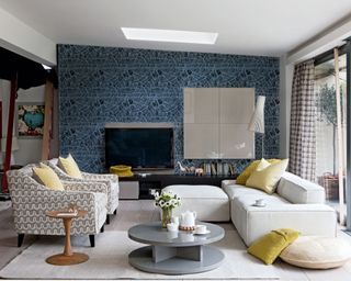 Open-plan family room space with large cream sofa and two cream patterned lounge chairs, accessorized with yellow cushions, light wooden flooring with cream rug, gray rounded coffee table and wooden rounded side table, feature wall with blue patterned wallpaper, gray storage cabinet mounted onto the wall, tv mounted to wall, low black sideboard