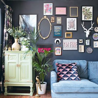grey living room wall decorated with gallery frames next to a mint green cupboard topped with flowers and a blue sofa with a patterned pillow