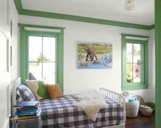 Children's bedroom with green painted ceiling and window trim, two windows, dark wooden flooring, single bed with checkered duvet, covered with cushions and soft toys, elephant painting on wall between windows