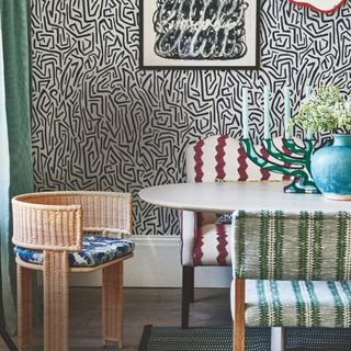 small dining room space with graphic wallpaper, artwork, white table, rattan chair with cushion, upholstered bench, patterned chair, patterned rug, decorative candelabra, blue vase