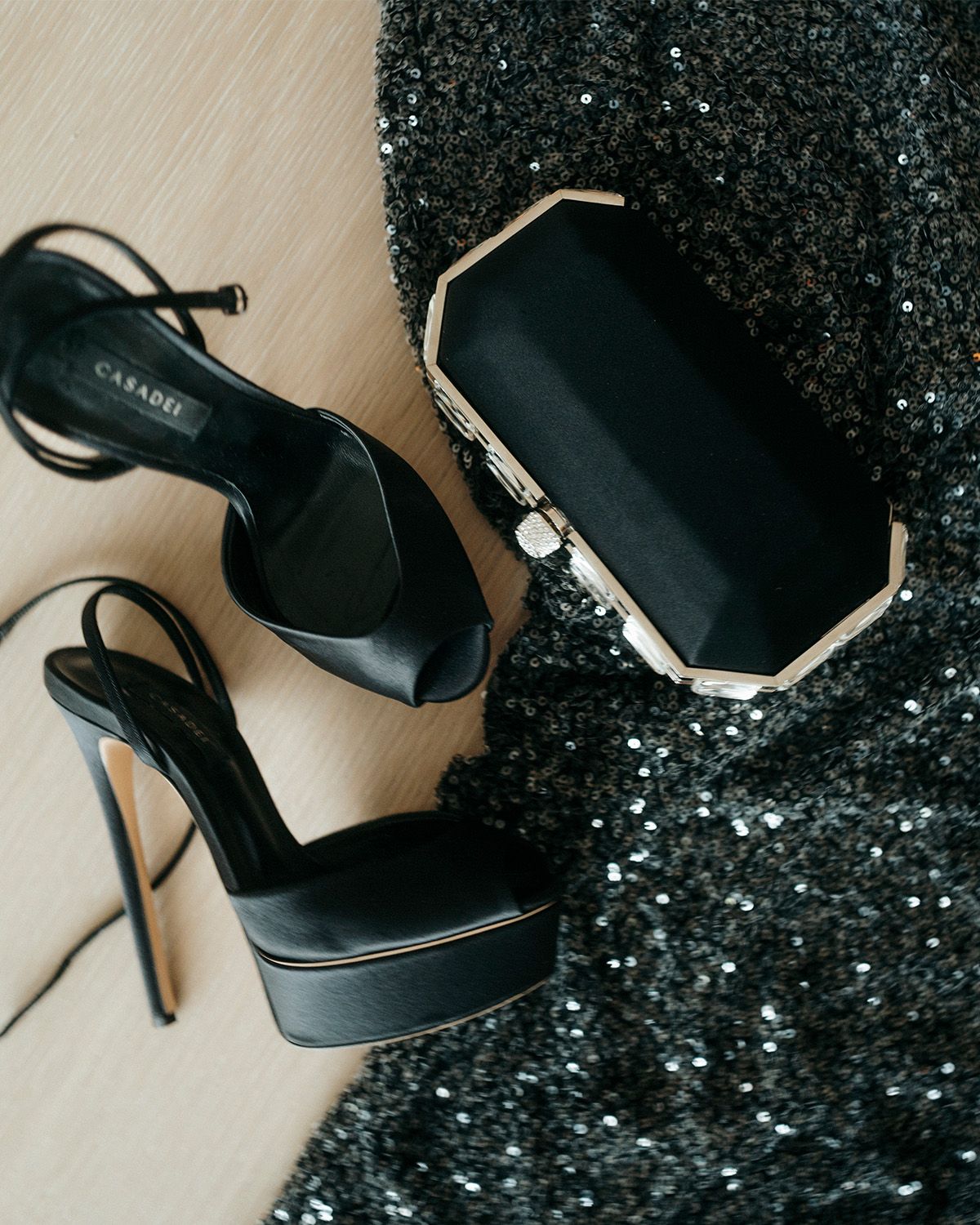Still life shot of Lydia Peckham's Casadei black stain peep-toe pumps, black clutch, and Patou black sequin dress.