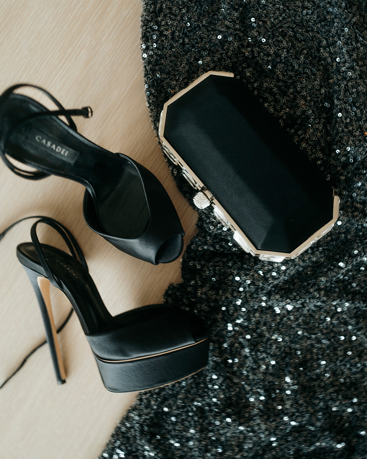 Still life shot of Lydia Peckham's Casadei black stain peep-toe pumps, black clutch, and Patou black sequin dress.