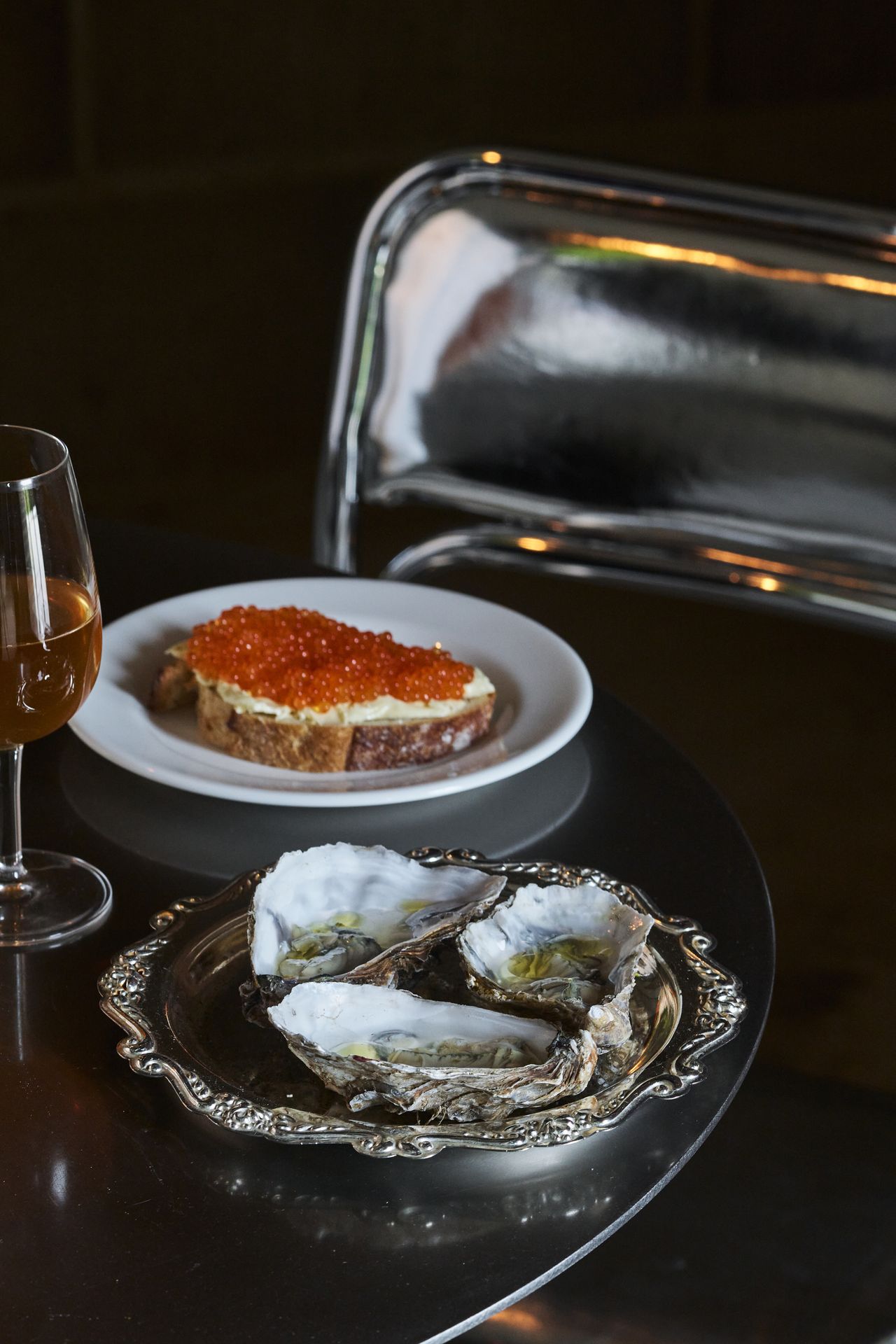A silvery plate with an ornate decoration is filled with three large-shell oysters and placed next to another white plate with a slice of bread, cheese, and orangey caviar and a orange wine glass in a chrome-clad restaurant.