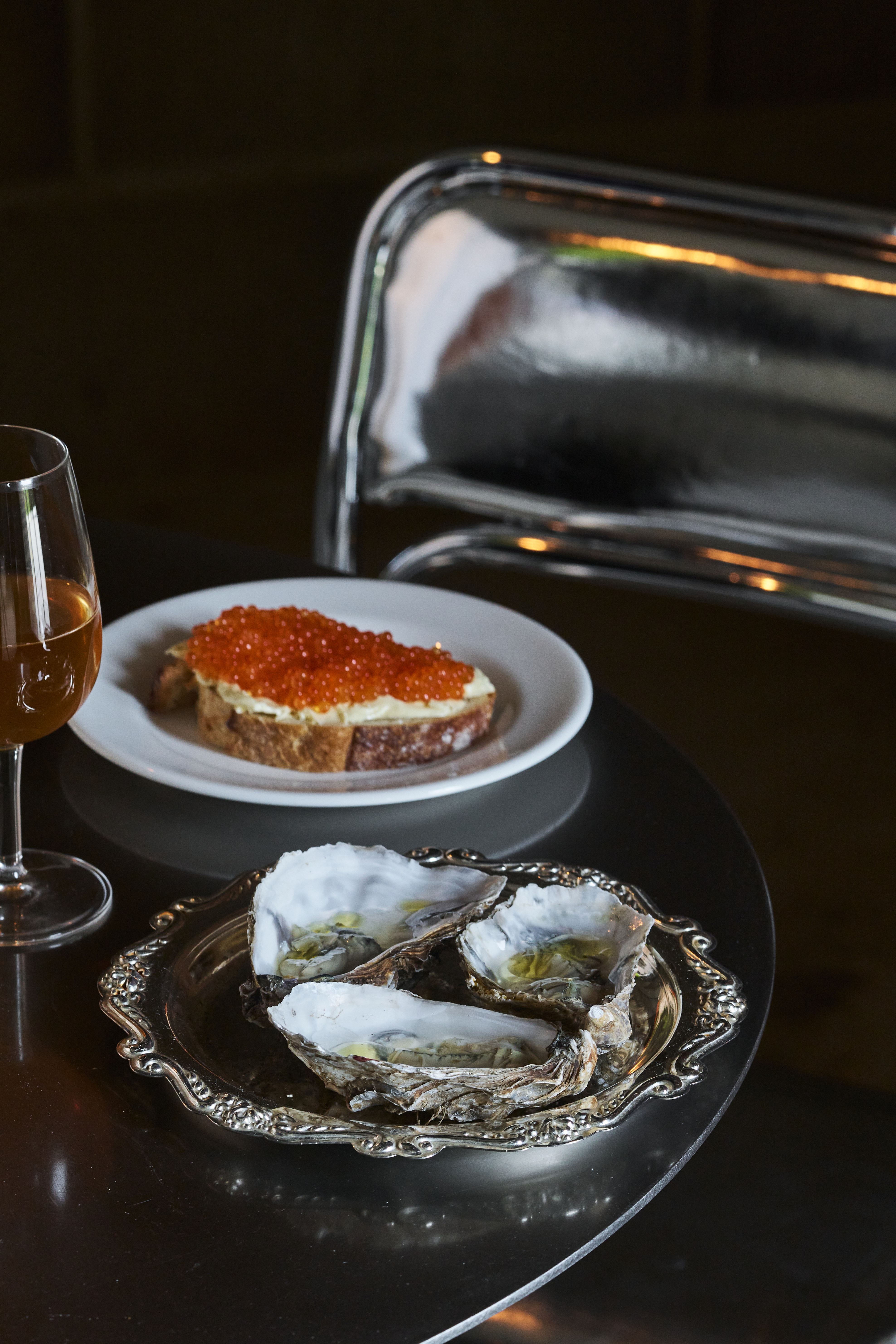 A silvery plate with an ornate decoration is filled with three large-shell oysters and placed next to another white plate with a slice of bread, cheese, and orangey caviar and a orange wine glass in a chrome-clad restaurant.
