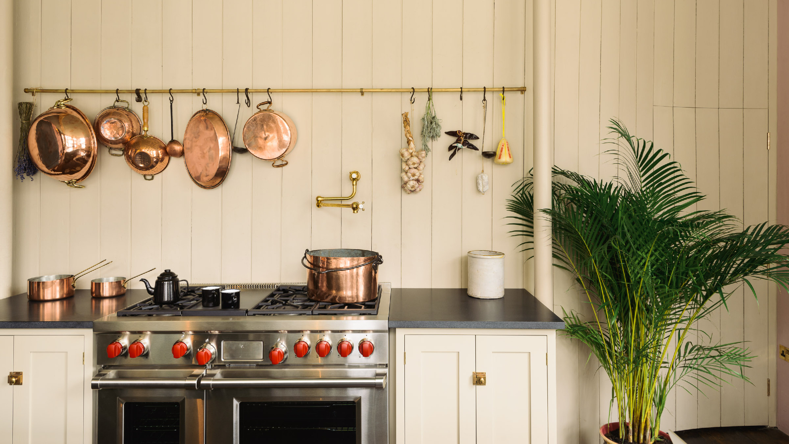 cream kitchen with shiplap walls, brass hanging rail with copper saucepans, black countertop, stainless steel stove with copper saucepan, plant in the right corner 
