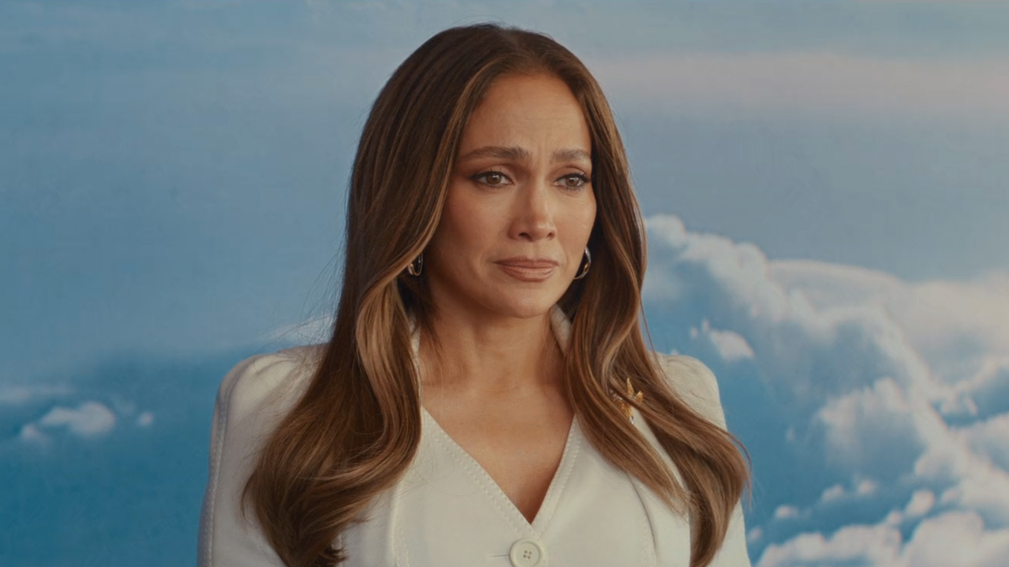 Jennifer Lopez wears a cream suit jacket with her hair blown out amidst a background of clouds in Office Romance.