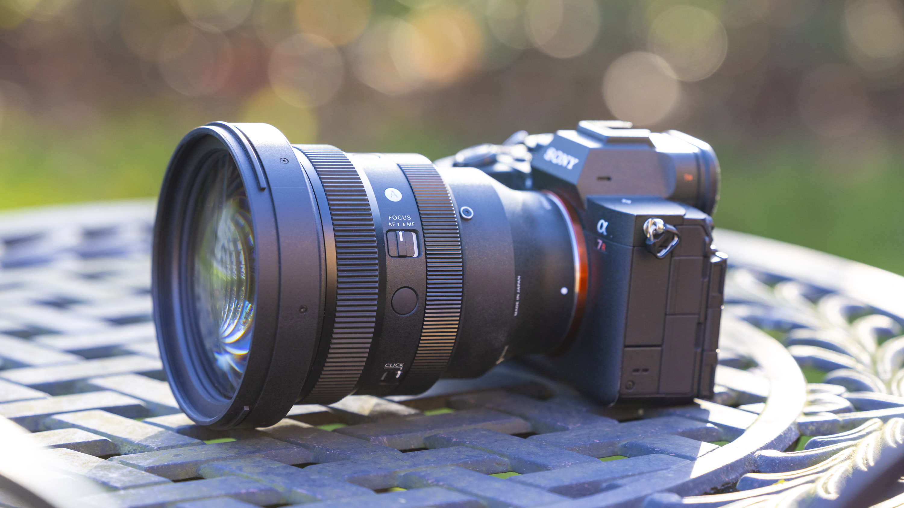 A side-on view of the Sigma 135mm f1.4 DG Art lens attached to a Sony A7R IV body on a metal garden table outdoors with lots of bokeh in the background