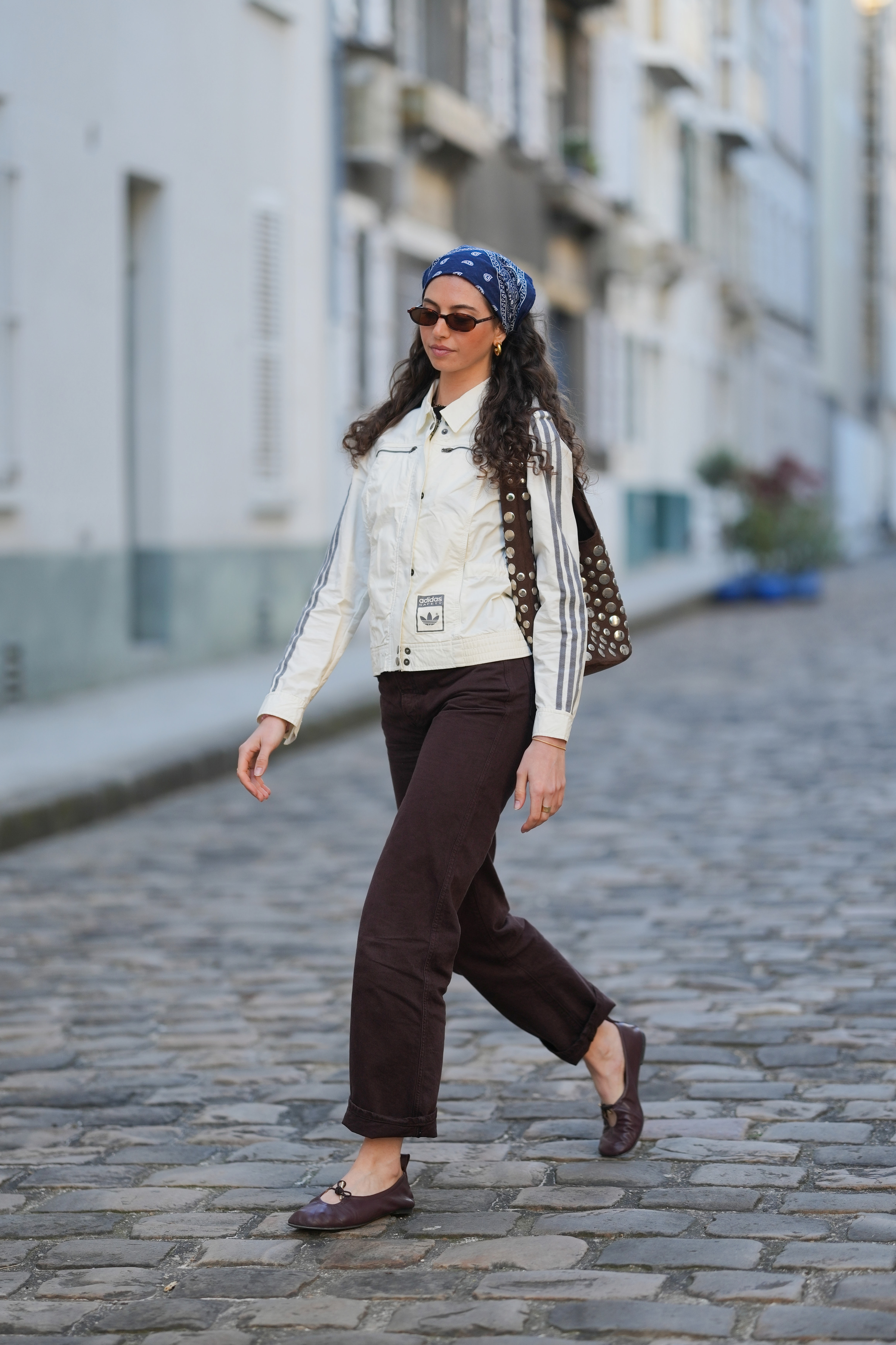 PARIS, FRANCE - APRIL 14: Amanda Derhy wears a blue scarf bandanna over the head with printed patterns, sunglasses, earrings, a white Adidas jacket with stripes on the sleeves, a bag with metal studs, brown denim pants / jeans with hem, brown / burgundy ballet flat shoes, during a street style fashion photo session, on April 14, 2026 in Paris, France. (Photo by Edward Berthelot/Getty Images)
