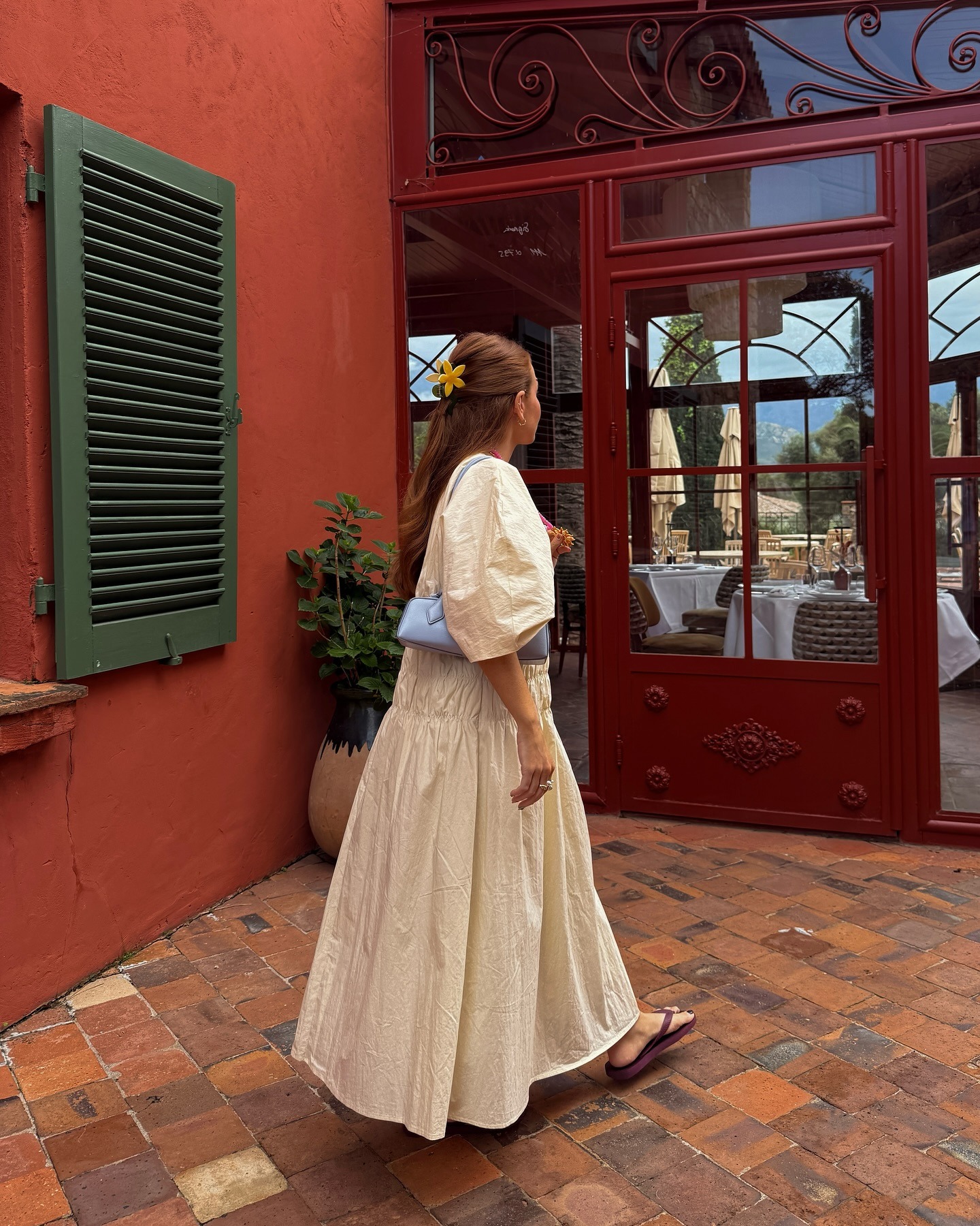 Fashion influencer Rebecca Ferraz Wyatt wears a butter yellow full length dress a pale blue Alaïa Le Teckel and burgundy flip flops in a recent image on holiday in Corsica.