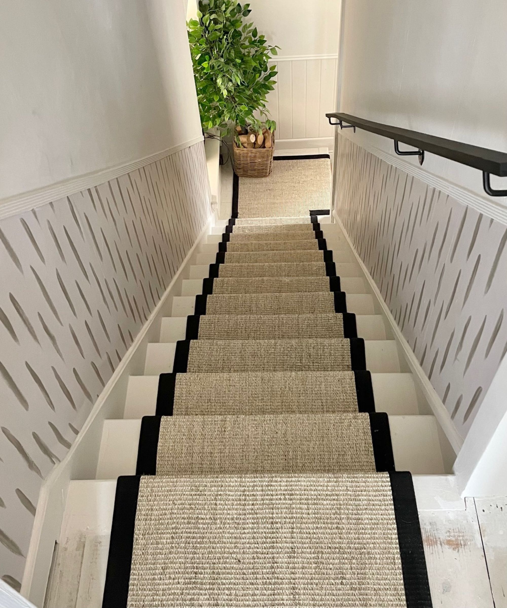 A downwards view of a beige woven stair runner with black edging on a white staircase with a black handrail and large plant at the bottom