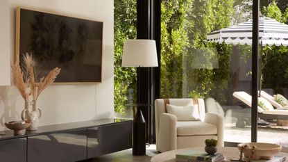 A beige chair in a living room with floor to ceiling window and flat screen TV hung on a wall