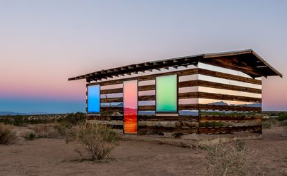 A wooden cabin in the desert with colourful mirrored strips running horizontally along the sides.