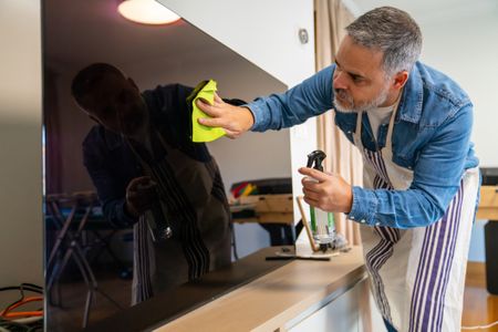 Adult man wearing an apron cleaning a large black tv screen with a spray bottle and microfiber cloth at home