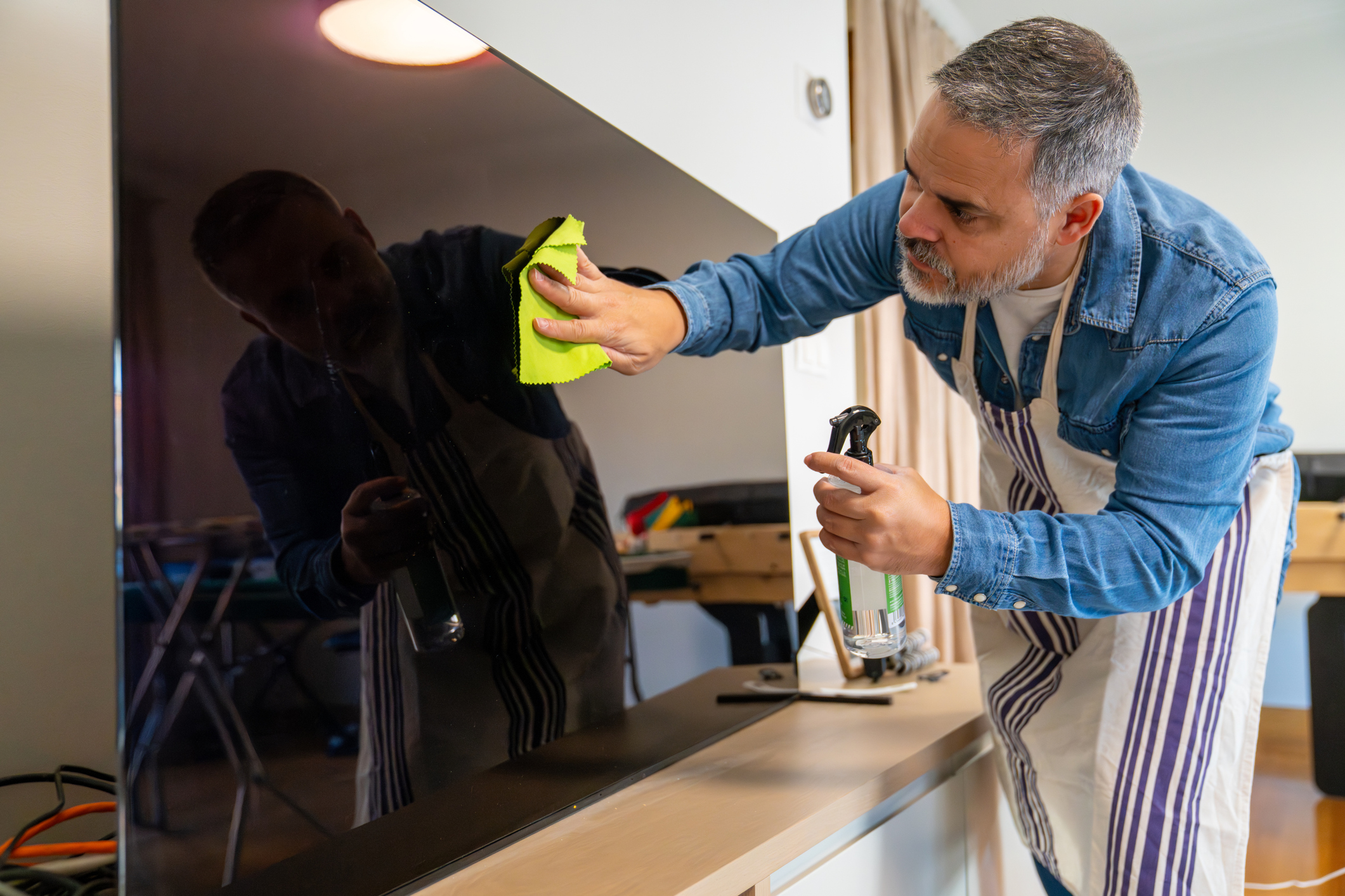 Adult man wearing an apron cleaning a large black tv screen with a spray bottle and microfiber cloth at home