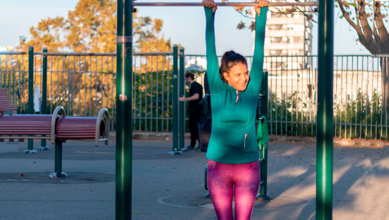 Woman hangs from a pull-up bar in a park. She is wearing pink leggings and a green long-sleeve athletic top.