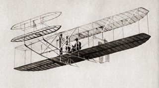 Historic black-and-white image of the Wright brothers' airplane in flight, showcasing their pioneering design with a biplane structure. The plane, with its exposed framework and fabric-covered wings, flies over Kitty Hawk, North Carolina.