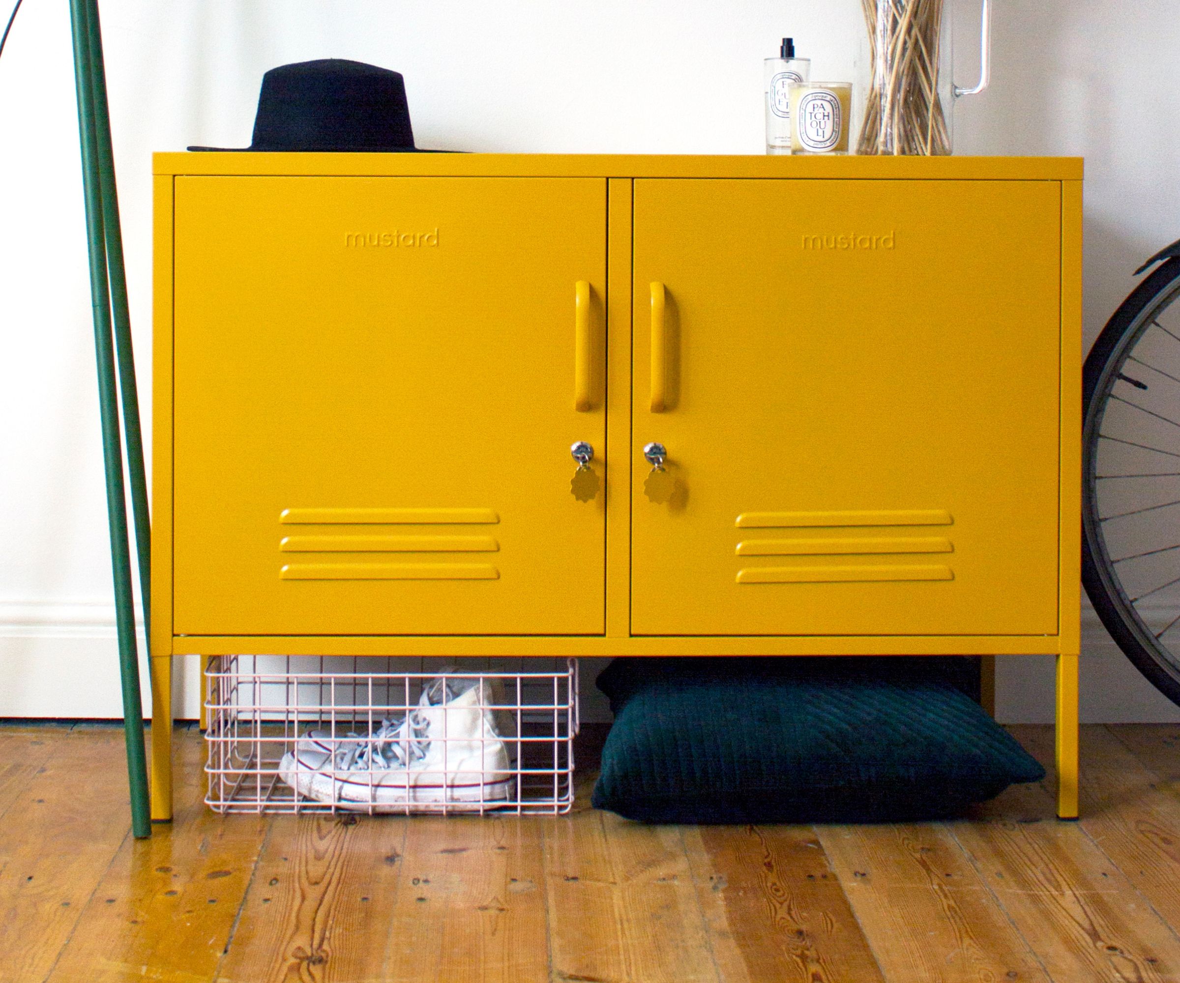 A bold mustard yellow powder-coated metal locker-style cabinet with two doors, louvred vents and key locks, branded "Mustard", sitting on short legs on bare wooden floorboards. A white wire basket with trainers and a dark cushion are stored underneath, with a black hat and fragrance bottle on top.