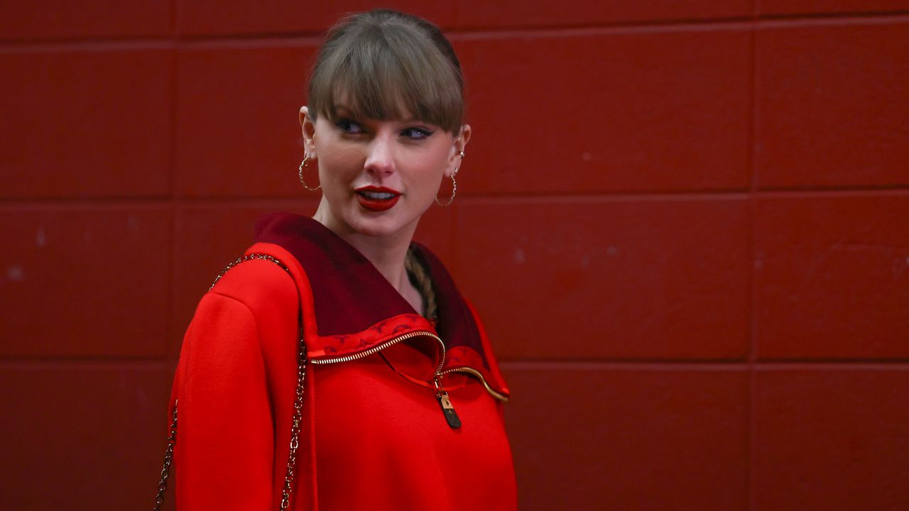 Taylor Swift wearing a red sweater standing in front of a red brick wall and smiling