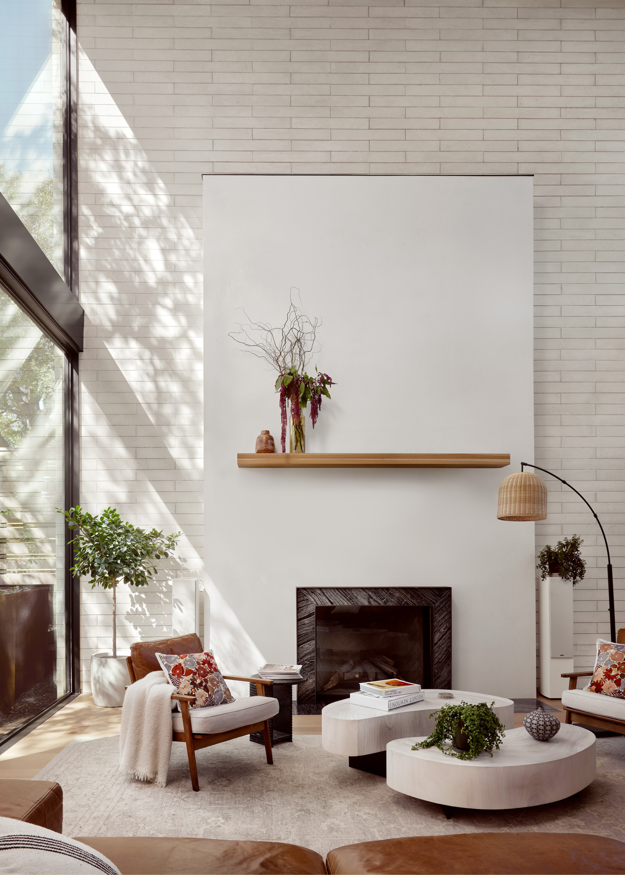 A white brick living room with a wooden floating shelf with a flower arrangement above a marble fireplace and a pair of coffee tables with wooden armchairs featuring patterned pillows and a throw