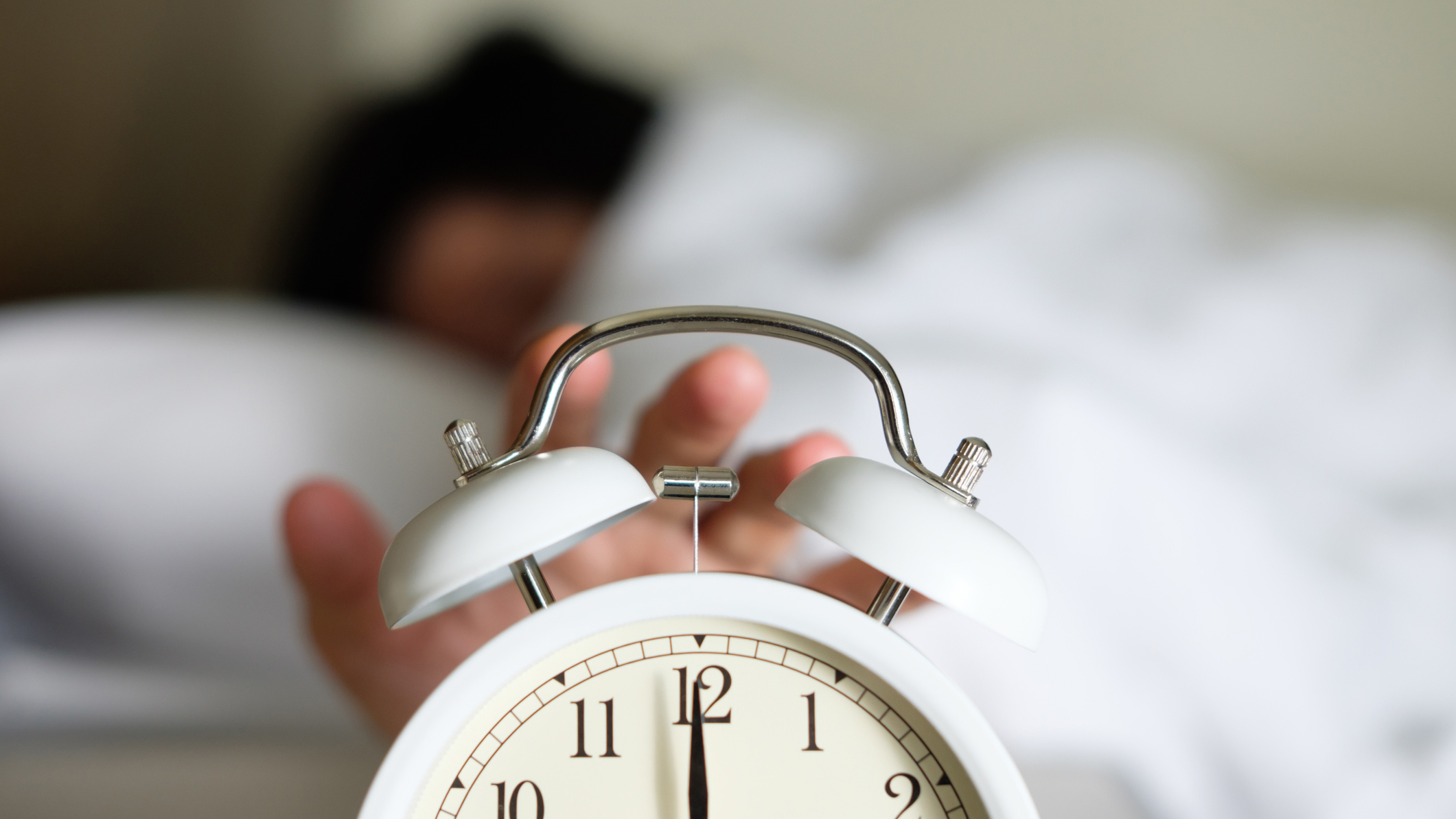 A woman with dark hair taps the top of a white alarm clock to stop it from ringing